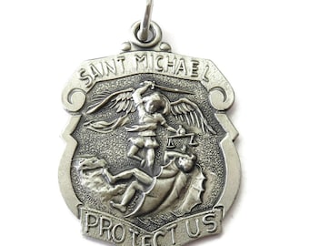 May include: Silver-toned pendant with the image of Saint Michael, featuring the text "Saint Michael Protect Us". The pendant depicts Saint Michael with wings, holding scales, and subduing a figure.