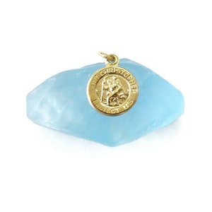 May include: A gold-colored Saint Christopher pendant with the words "Saint Christopher Protect Us" encircling an image of Saint Christopher. The pendant is round and hangs from a small loop. It rests on a light blue, irregularly shaped stone.