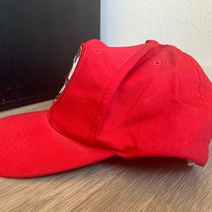 May include: A red baseball cap with a black and white patch on the front. The patch features a stylized image of a person's face.