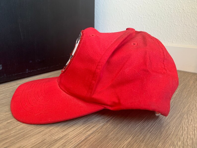 May include: A red baseball cap with a black and white patch on the front. The patch features a stylized image of a person's face.