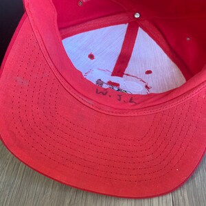 May include: Red baseball cap with a white interior and a small embroidered logo. The inside of the cap has a white tag with the text "W.J.L."