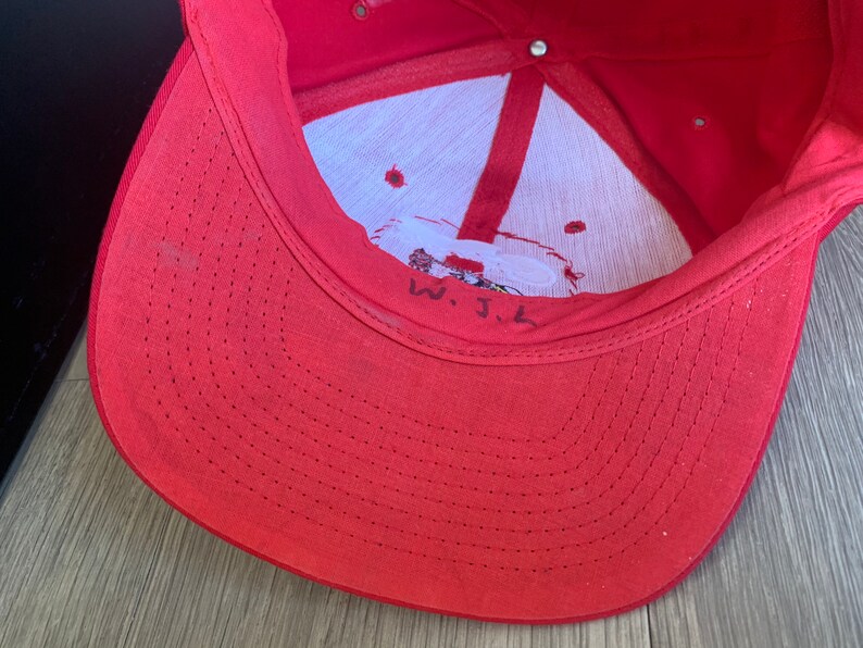 May include: Red baseball cap with a white interior and a small embroidered logo. The inside of the cap has a white tag with the text "W.J.L."