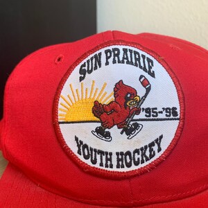 May include: Red baseball cap with a white patch featuring a cardinal, a hockey stick, a sunrise, and the text "Sun Prairie Youth Hockey '95-'96".