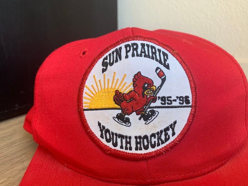 May include: Red baseball cap with a white patch featuring a cardinal, a hockey stick, a sunrise, and the text "Sun Prairie Youth Hockey '95-'96".