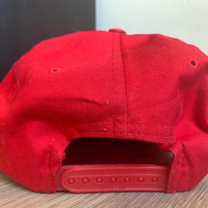 May include: A red baseball cap with a black adjustable strap. The strap has six holes for size adjustment.