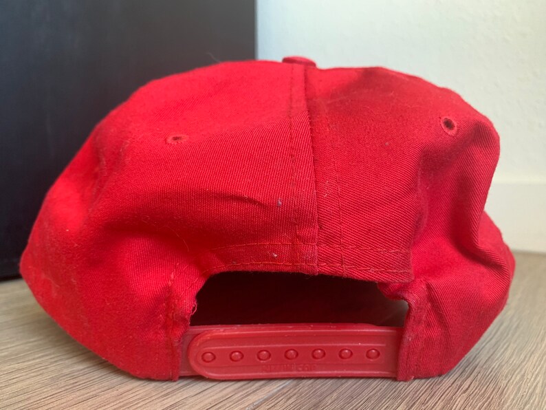 May include: A red baseball cap with a black adjustable strap. The strap has six holes for size adjustment.