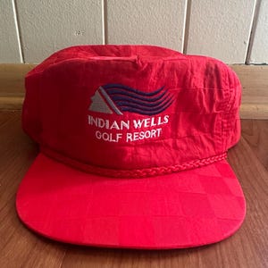 May include: A red golf cap with a checkered brim. The cap features the embroidered text "INDIAN WELLS GOLF RESORT" in white, along with a blue and white graphic. The cap has a braided trim.