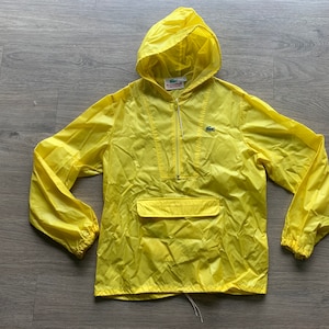 May include: A yellow hooded raincoat with a zipper closure and a small pocket on the front. The raincoat has a green crocodile logo on the chest and the text "Lacoste" on the zipper pull.