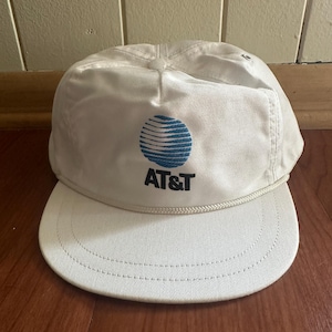 May include: A white AT&T baseball cap with a curved brim and a rope detail. The cap features the AT&T logo, a blue globe design, and the AT&T text in black. The cap is made of a durable fabric.