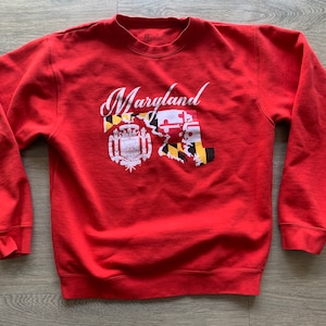 May include: Red sweatshirt with a white graphic of the state of Maryland with the state flag and the word "Maryland" in a white cursive font.
