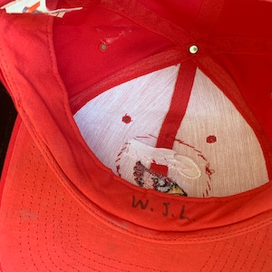 May include: Red baseball cap with a white mesh lining and a small embroidered logo. The inside of the cap has handwritten text that says "W.J.L."