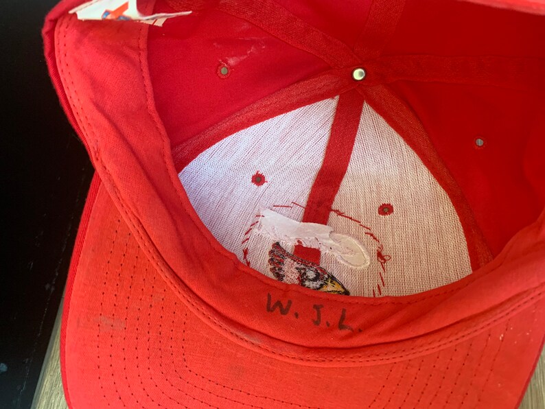 May include: Red baseball cap with a white mesh lining and a small embroidered logo. The inside of the cap has handwritten text that says "W.J.L."