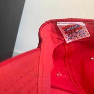 May include: Red baseball cap with a white label that reads "NISSIN WL MADE IN PHILIPPINES NISSIN CAP".