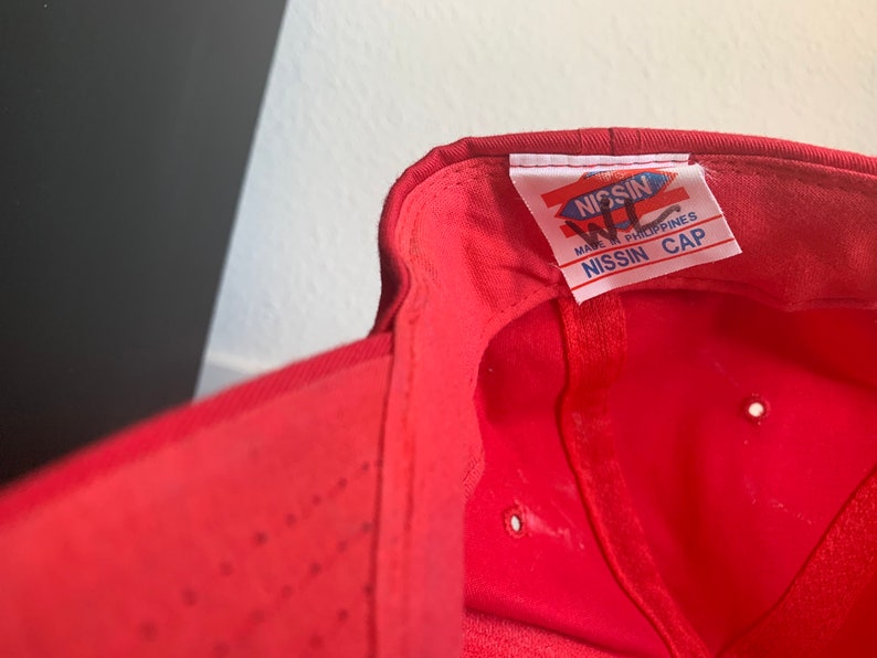May include: Red baseball cap with a white label that reads "NISSIN WL MADE IN PHILIPPINES NISSIN CAP".