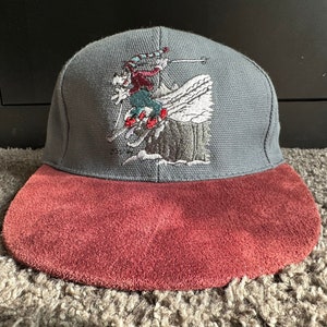 May include: A grey and burgundy baseball cap with an embroidered design of a person skiing down a mountain. The cap has a suede brim.