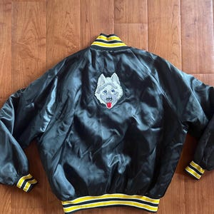 Pode incluir: Casaco bomber de cetim preto com um patch de cão husky branco e cinzento nas costas. O casaco tem acabamentos listrados amarelos e brancos na gola, punhos e bainha. Estilo universitário clássico.