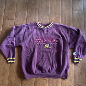 May include: A vintage purple sweatshirt with the word "Minnesota" in script across the chest, along with a graphic of a mascot. The collar and cuffs have yellow and gold stripes. The Starter logo is visible on the sleeve.