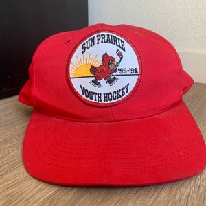 May include: Red baseball cap with a circular patch featuring a cartoon cardinal holding a hockey stick. The patch reads "Sun Prairie Youth Hockey '95-'96".