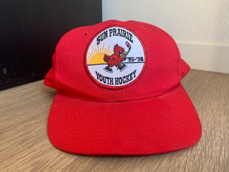 May include: Red baseball cap with a circular patch featuring a cartoon cardinal holding a hockey stick. The patch reads "Sun Prairie Youth Hockey '95-'96".