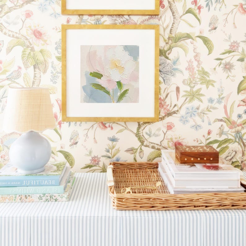 May include: A light blue ceramic table lamp with a scalloped shade sits on a stack of books. Next to it, a rectangular woven wicker tray holds a stack of books and a small wooden box with gold inlay, all against a floral wallpaper background.
