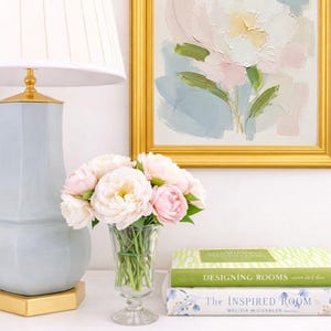 May include: A light blue table lamp with a white pleated shade, a clear glass vase filled with pale pink peonies, and two stacked books titled Designing Rooms and The Inspired Room, next to a framed abstract floral painting in a gold frame.