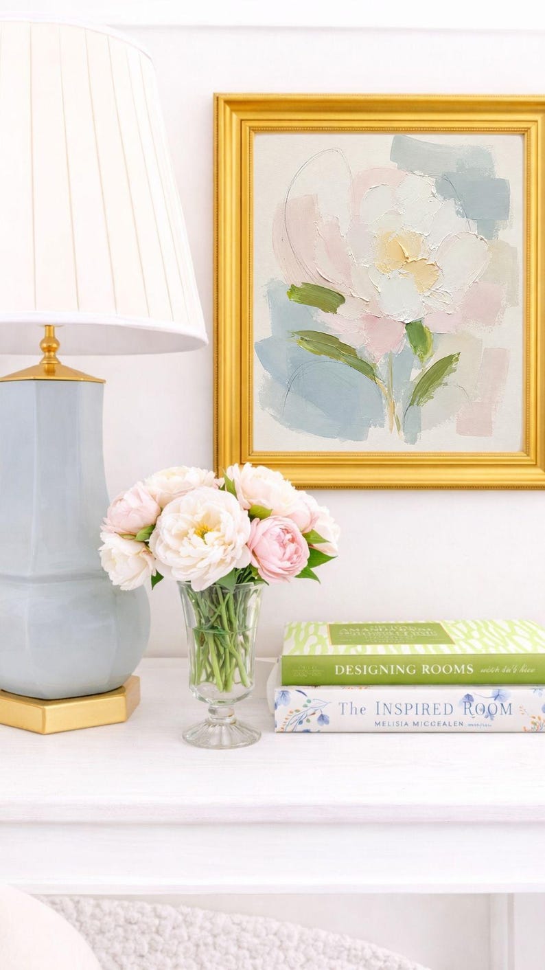 May include: A light blue table lamp with a white pleated shade, a clear glass vase filled with pale pink peonies, and two stacked books titled Designing Rooms and The Inspired Room, next to a framed abstract floral painting in a gold frame.