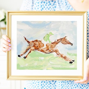 A person wearing a blue patterned shirt holds a framed painting of a galloping horse.
