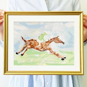 A person holds a gold-framed painting of a brown horse and jockey running across a green field against a blue sky.