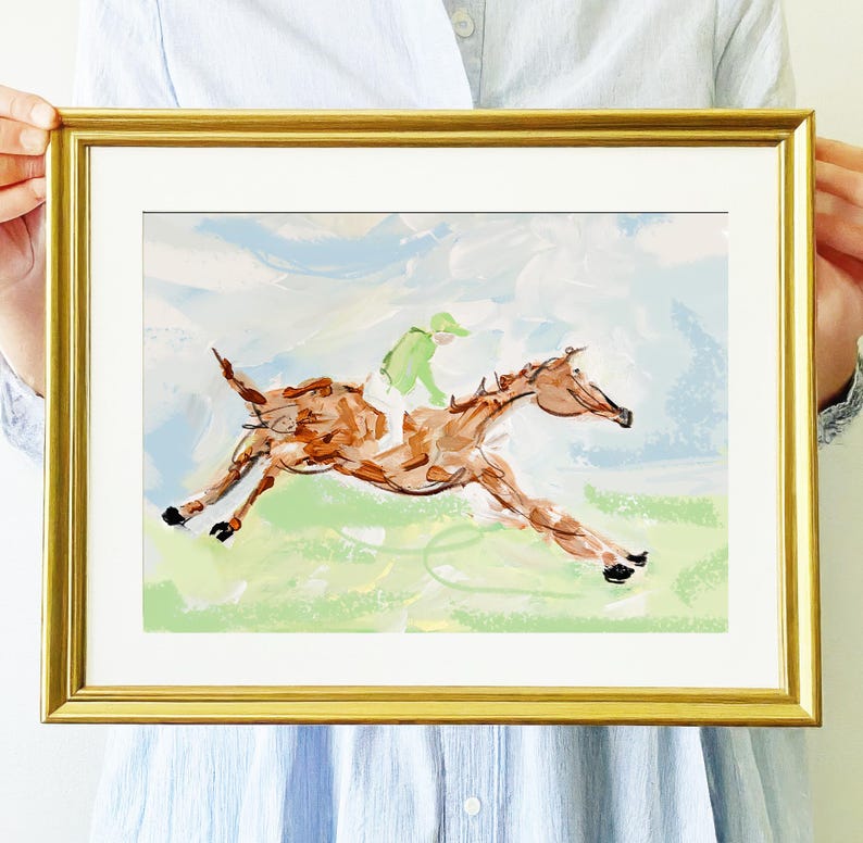 A person holds a gold-framed painting of a brown horse and jockey running across a green field against a blue sky.