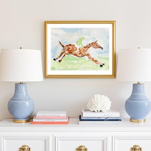 A gold-framed painting of a racing horse centered on a wall between two lamps with blue ceramic bases and white shades.