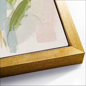 May include: A framed canvas print featuring abstract brushstrokes in shades of sage green, soft blue, and pale pink on a white background. The artwork is encased in a metallic gold frame with a textured finish, viewed from an angled perspective.
