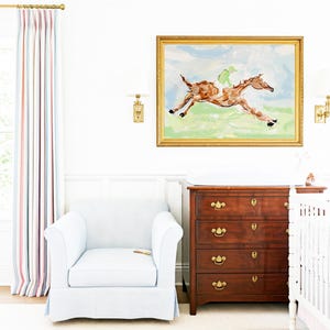 A nursery interior shows the framed horse painting mounted on a wall above a dark wood dresser, next to a light blue armchair and striped curtains.