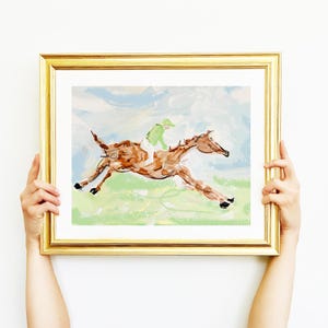 A person's hands hold a framed impressionist horse painting against a plain white background.