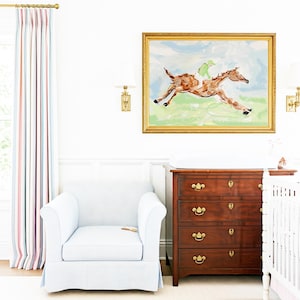 A nursery features the framed horse artwork displayed on a white wall above a wooden dresser and a light blue upholstered chair.