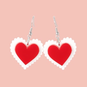 May include: A pair of red heart-shaped earrings with a white scalloped edge. The earrings are made of acrylic and have silver hooks.