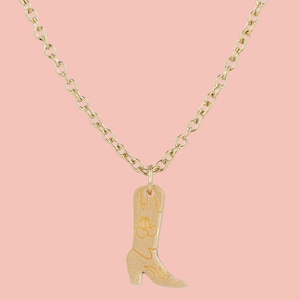 May include: Gold-coloured necklace featuring a cowboy boot pendant. The boot pendant has floral and swirl designs. The chain and pendant are gold-toned, set against a light pink background.
