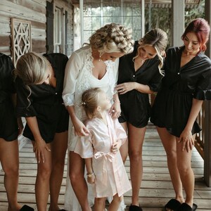 May include: A group of women in black satin robes and a bride in a white lace robe stand together. A young girl in a pink satin robe stands in front of the bride. The women are smiling and looking at the camera.