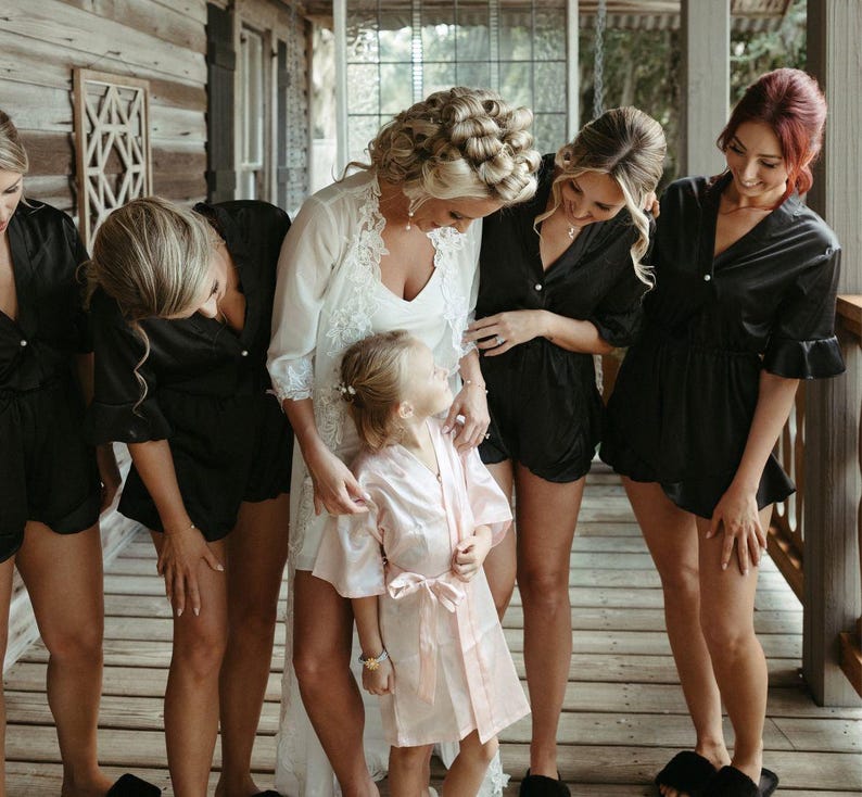 May include: A group of women in black satin robes and a bride in a white lace robe stand together. A young girl in a pink satin robe stands in front of the bride. The women are smiling and looking at the camera.