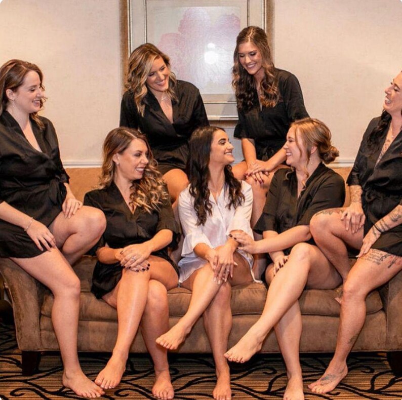 May include: A group of six women wearing black robes and a white robe are sitting on a brown couch. They are all smiling and looking at each other. The woman in the white robe is sitting in the center of the couch.