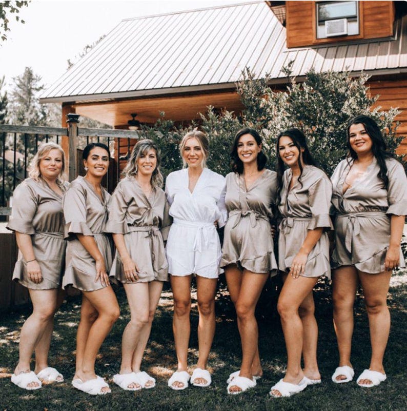 May include: Seven women wearing matching satin robes in a light brown color. The robes have long sleeves and a tie at the waist. The women are standing in a row, smiling at the camera. They are wearing white fluffy slippers.