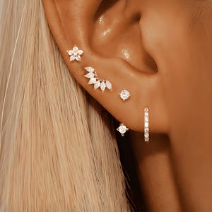 May include: A close-up of a person's ear with multiple ear piercings. The piercings feature small, sparkly, clear gemstones set in gold. The ear is pierced in the helix, conch, and lobe.