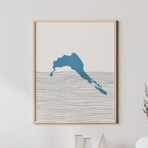 Op de afbeelding: Een ingelijste kunstdruk met een blauwe abstracte vorm boven horizontale lijnen, die een landschap suggereren. Het kunstwerk hangt boven een licht houten kast met een geweven voorkant, een witte vaas en een houten stoel.