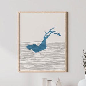 Pode incluir: Uma impressão abstrata emoldurada com uma forma azul que se assemelha a uma onda ou uma ilha sobre um fundo branco com linhas pretas finas. A impressão está pendurada em uma parede branca acima de um armário de madeira clara com uma frente de vime tecida.