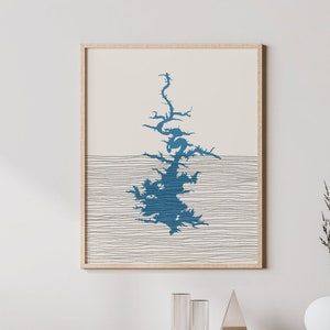 May include: A minimalist art print featuring a blue lake outline on a cream background with thin black lines. The print is framed in a light wood frame and hangs on a white wall above a wooden cabinet.