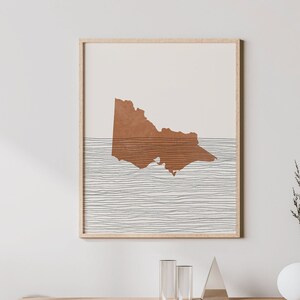 May include: A minimalist art print featuring a brown outline of the state of Victoria, Australia, against a white background with thin black lines. The print is framed and hanging on a white wall above a wooden cabinet.