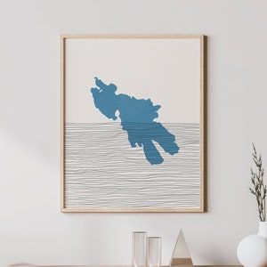 Puede incluir: Una impresión enmarcada de una forma abstracta azul sobre un fondo blanco con finas líneas negras. La impresión está colgada en una pared blanca sobre un armario de madera clara con un frente de ratán tejido.