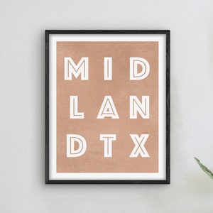 May include: A framed print with a peach background and white text that reads "MID LAND DTX". The print is hanging on a white wall with a clock and a lamp on a white table.