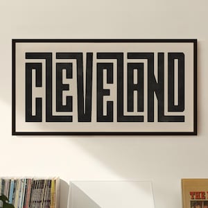 May include: A black and white framed print with the word "CLEVELAND" in a geometric font. The print is hanging on a white wall above a record player and a shelf of vinyl records.