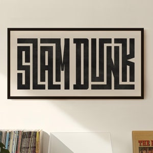 May include: A black and white framed print with the text "SLAM DUNK" in a bold, geometric font. The print is hanging on a white wall above a wooden cabinet with a record player and a stack of vinyl records.