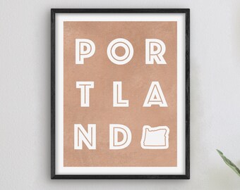 May include: A framed print with a brown background and white text that reads "PORTLAND" with an outline of the state of Oregon. The print is hanging on a white wall with a clock and a lamp on a white table.